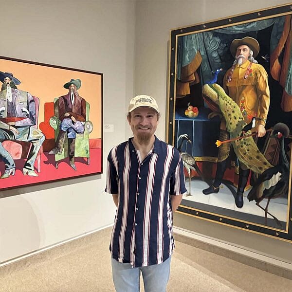 Poet and actor Evan Wambeke stands in the Whitney Western Art Museum with two paintings featuring interpretations of Buffalo Bill. Wambeke gave a poetry reading and workshop in the summer of 2024 at the Whitney Western Art Museum as part of an artist in residency program.
