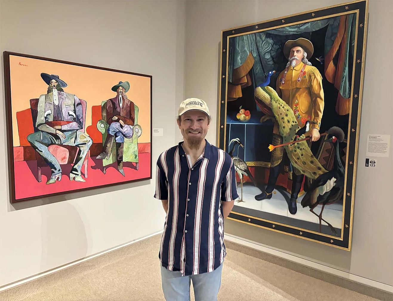 Poet and actor Evan Wambeke stands in the Whitney Western Art Museum with two paintings featuring interpretations of Buffalo Bill. Wambeke gave a poetry reading and workshop in the summer of 2024 at the Whitney Western Art Museum as part of an artist in residency program.
