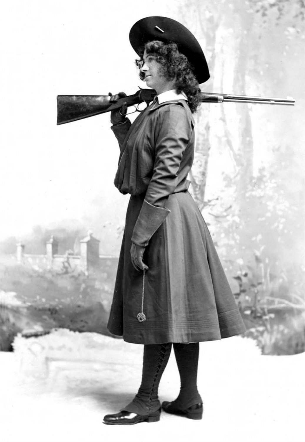 Woman in dress and wide-brimmed hat standing to side with rifle over shoulder.