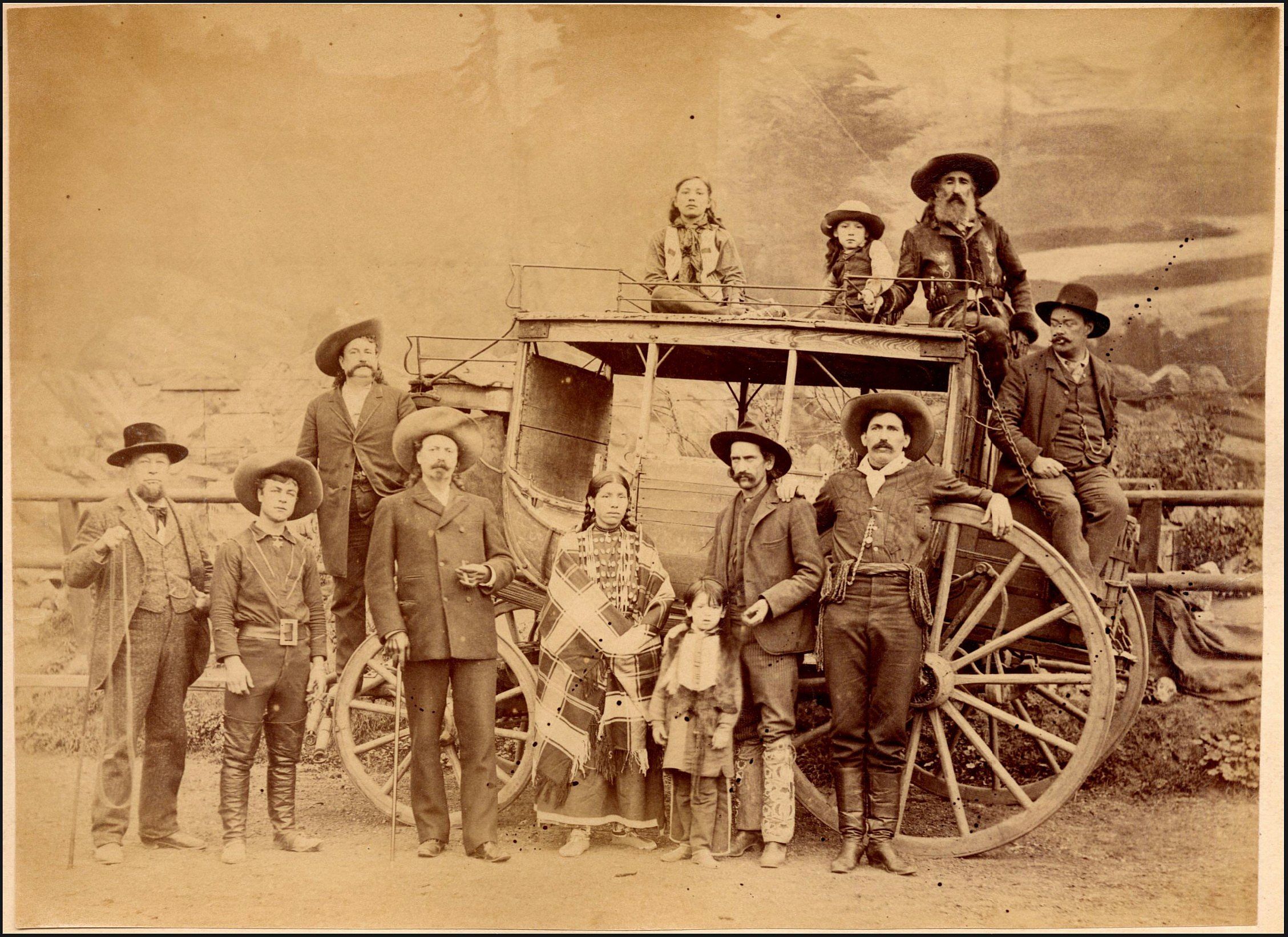 William F. Cody and the Deadwood Stagecoach 1884; A photograph of Unknown Man, Johnny Baker, William F. Cody, John Burke, William "Broncho Bill" Irving, Irving's wife Ella and son Bennie, Buck Taylor, two native boys, John Nelson, and unknown man on and around the Deadwood stagecoach parked in front of a rail fence. Rocks, vegetation and a painted backdrop of mountains and river is in the background. P.69.2041.1