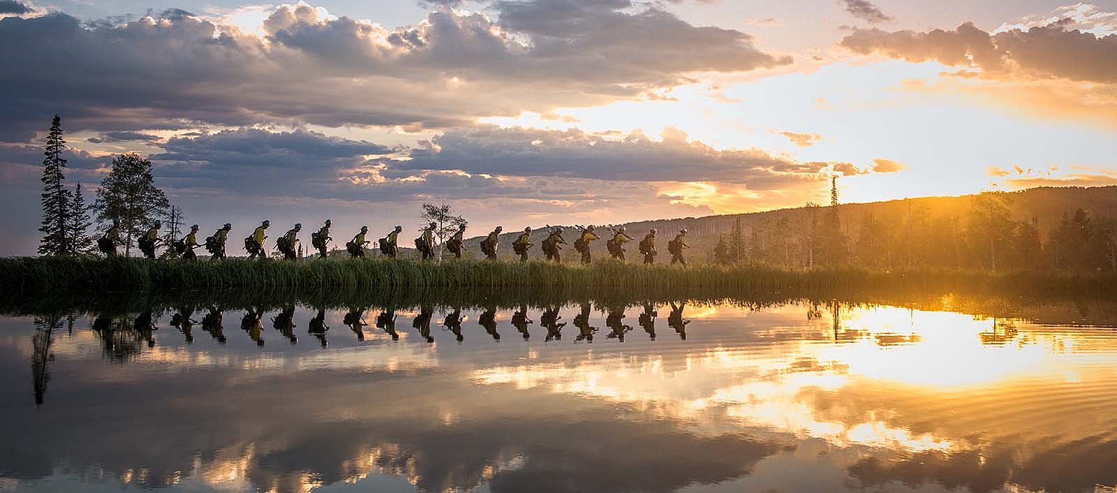 Line of firefighters walking along water at sunrise or sunset. "Reflections," Fire on the Mountain: Photographs of Wildland Firefighting by Kyle Miller. (detail)