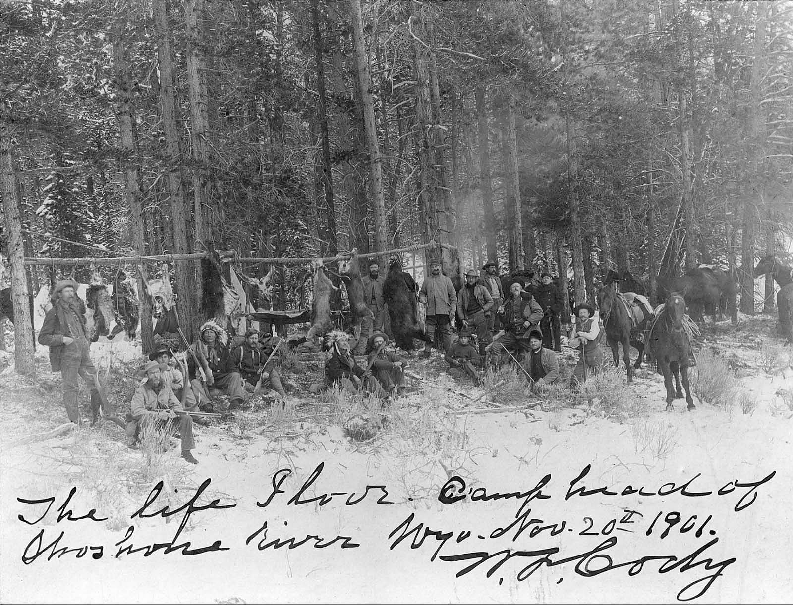 William F. Cody, Chief Iron Tail, and Mike Russell (lower left front) with a hunting party of Native American and European American men holding rifles in a snow covered wooded area. Handwritten caption, "The life I love. Camp head of Shoshone river Wyo. Nov. 20th 1901. W. F. Cody." MS 6 William F. Cody Collection. P.69.1062