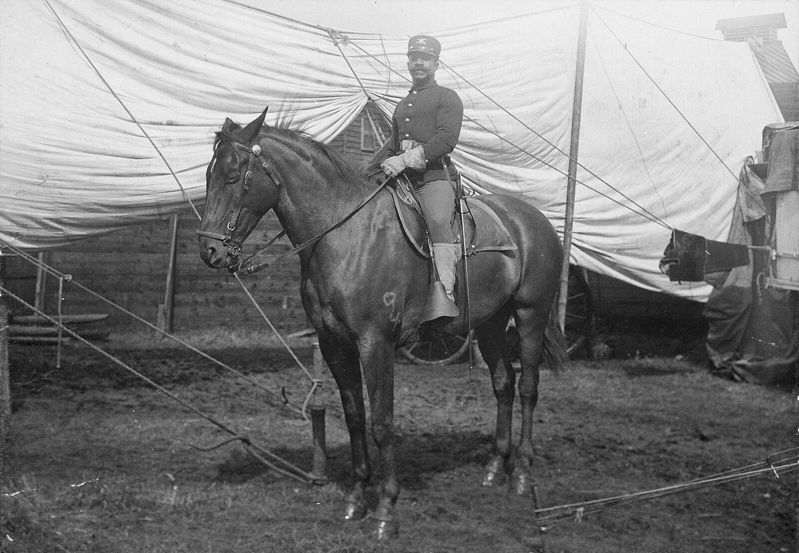 An African American man mounted on a dark horse in front of show tent in back lot of wild west show.
