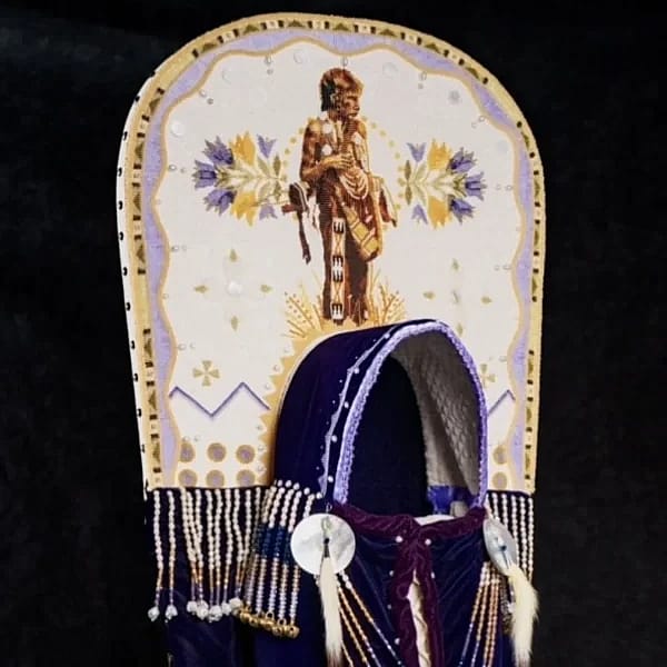 A Blackfeet story of the prairie crocus cradleboard by artist Jackie Larson Bread (Blackfeet). The cradleboard features tanned hide, velveteen, glass beads, shell, ermine and copper bells.