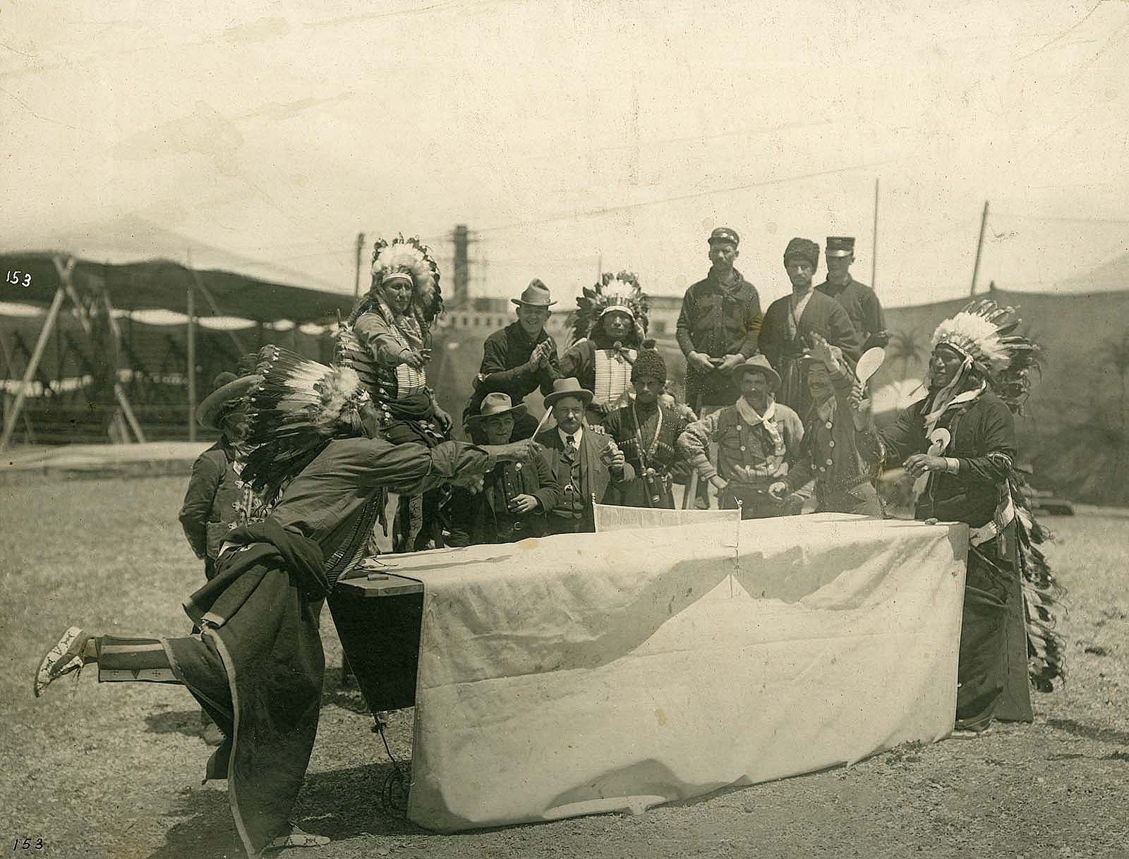 A performer who may be Oglala Lakota Chief Iron Tail plays ping pong with a fellow castmate as Joe Black Fox and a group Native performers, cowboys, vaqueros, and Cossacks watch the match. MS 6 William F. Cody Collection, McCracken Research Library. P.69.1031