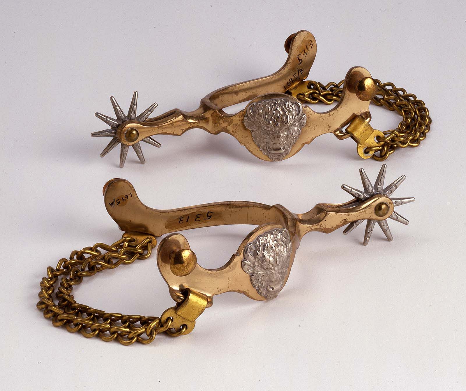 Pair of spurs, brass with nickel-silver buffalo heads and chain under shanks. 1.69.5